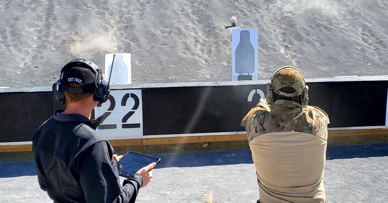 Two shooters on an outdoor qualification line engaging white silhouette targets, one of them holding a control tablet.