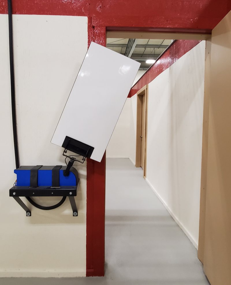 A lift unit bolted to a shoot-house wall, swinging a white target out across a doorway into the corridor beyond.