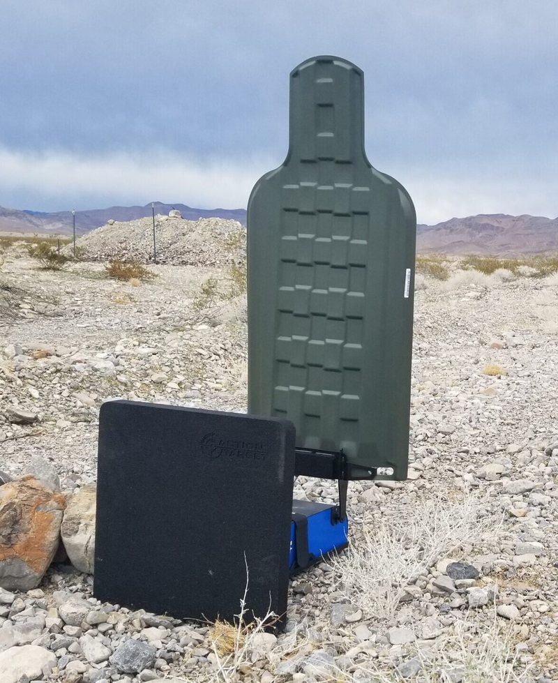 A green plastic E-Type silhouette raised on an AutoTargets lift unit standing on open desert ground behind a black rubber block ballistic stand.