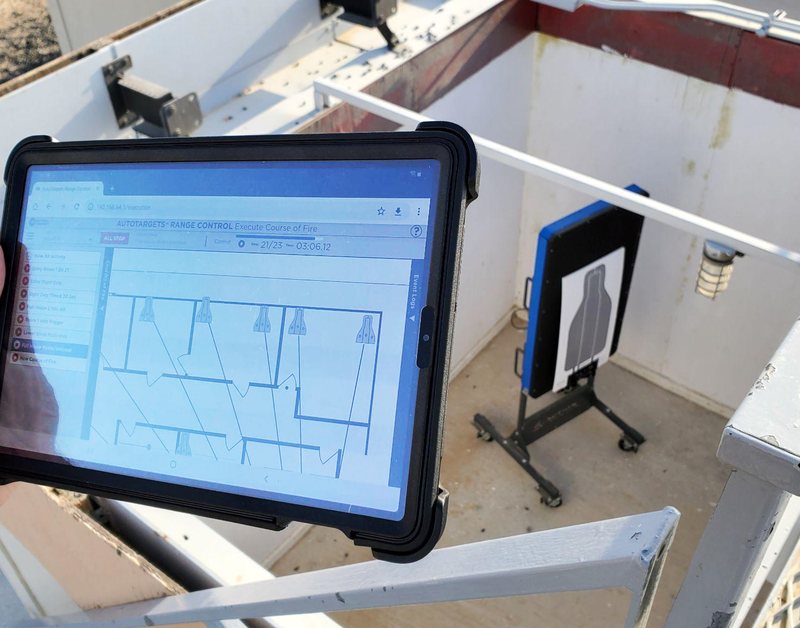 A hand holding a ruggedized tablet running the Range Control course-of-fire execution screen, with a silhouette target on a lift unit in the background.