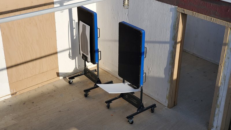 Two wheeled portable bullet traps with blue-framed steel plates standing in a shoot-house bay, the nearer one carrying a paper silhouette.